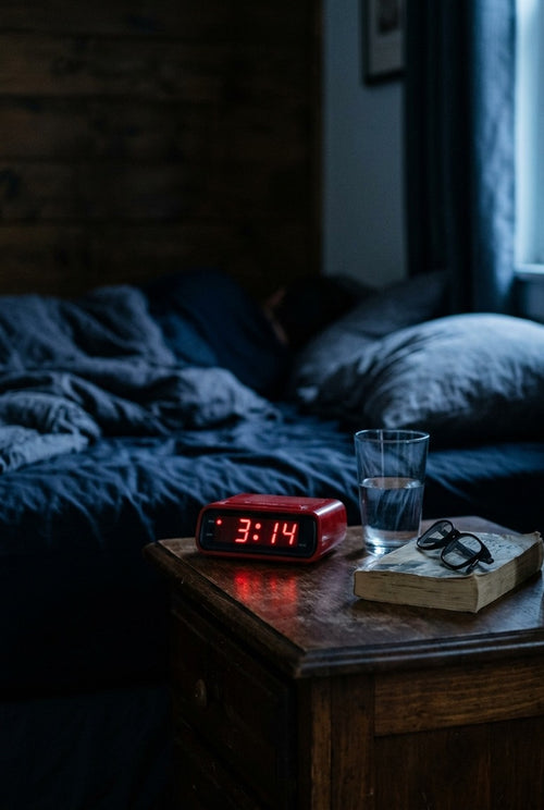 A dark bedroom at 3:14 a.m.: a red alarm clock, a glass of water and reading glasses on a book beside an unmade bed